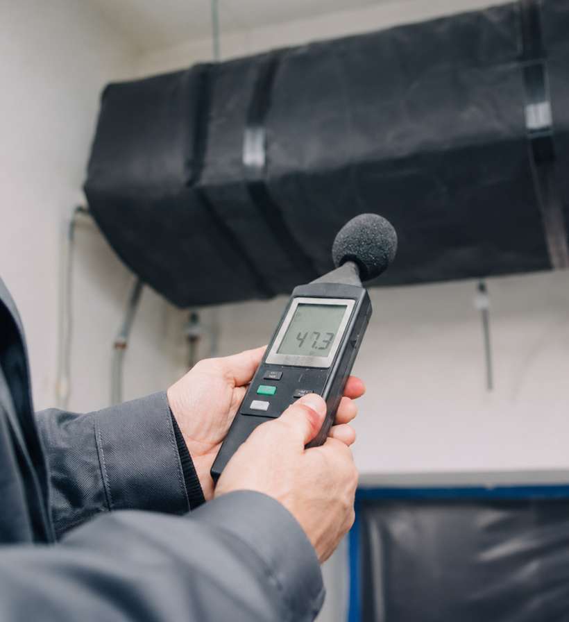 Professional acoustic measurement on a black insulated HVAC duct using a sound level meter for building noise control.