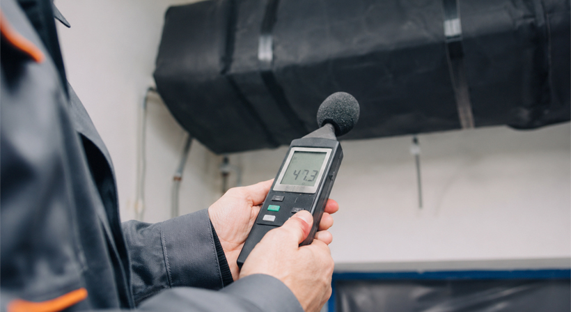Professional acoustic measurement on a black insulated HVAC duct using a sound level meter for building noise control.
