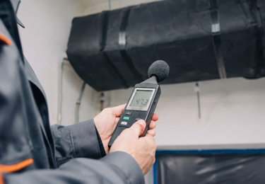 Professional acoustic measurement on a black insulated HVAC duct using a sound level meter for building noise control.