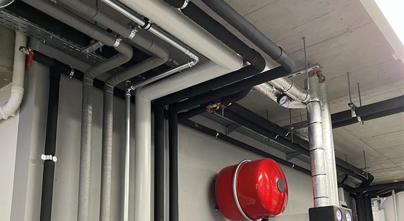 Mechanical room with insulated pipes, control equipment and a red wall-mounted component.